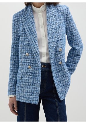Vrouw draagt een blauwe dubbelgeknoopte tweed blazer met zilveren knopen, een witte blouse met gerimpelde kraag en donkerblauwe hoge taille jeans.