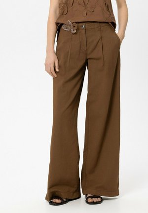 Braune Hosen mit weitem Bein und vorderen Falten, einer dekorativen Stoffblume am Bund, kombiniert mit schwarzen offenen Sandalen und einem passenden braunen Oberteil.