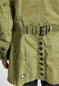 Olivgrön jacka med bälte i midjan, med knäppning fram, framträdande sömmar och en lapp med en logga på nedre vänstra sidan.