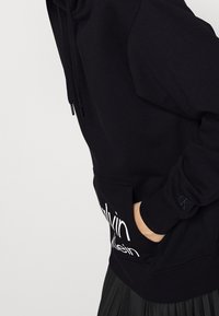 Osoba v černé mikině Calvin Klein s kapucí, ruka v přední kapse, viditelné logo CK na manžetě a čern�é plisované sukni pod tím.