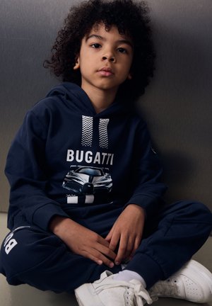 Bambino piccolo con capelli ricci che indossa una felpa con cappuccio Bugatti blu navy e pantaloni abbinati, seduto a gambe incrociate su sfondo grigio.