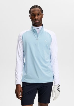 Mężczyzna ubrany w jasnoniebieską i białą golfową koszulę z długim rękawem, czarne spodenki i białą rękawiczkę golfową, stojący na jednolitym jasnym tle.