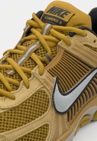 Baskets Nike Vomero 5 jaunes avec accents gris, présentant des panneaux en maille, des superpositions texturées et un logo Swoosh argenté sur le côté.