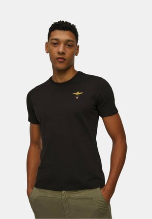 Camiseta negra de manga corta hecha de algodón suave, con un pequeño emblema dorado en el pecho y un escote redondo.