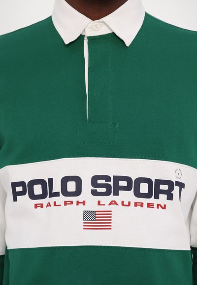 Grön polo med vit krage, med en framträdande "POLO SPORT"-logotyp i marinblått och rött, samt en amerikansk flagga som accent. Strukturerad bomullsmaterial.