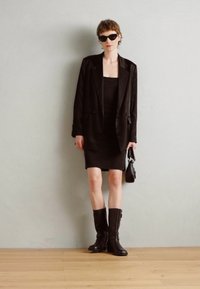 Černý blazer přes přiléhavé černé šaty, doplněný černými kozačkami do kolen a malou kabelkou. Hladká tkanina, strukturovaný design a minimální akcenty.
