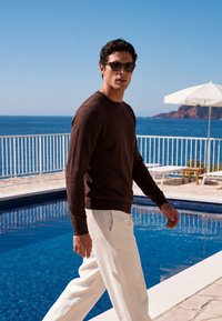 Homme portant des lunettes de soleil, un pull marron et un pantalon blanc marche à côté d'une piscine avec vue sur l'océan et les falaises sous un ciel bleu dégagé.