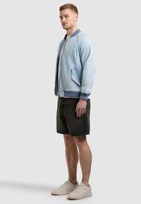 Jeune homme debout portant une veste bomber bleu clair, un t-shirt blanc, un short noir jusqu'aux genoux et des baskets blanches, devant un fond uni.