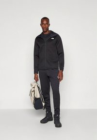 Zwarte jas met ritssluiting en een gestructureerd oppervlak, gecombineerd met zwarte broek en stevige schoenen. De persoon draagt een beige en marineblauwe rugzak.