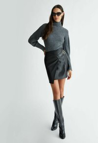 Maglione a collo alto grigio a costine, gonna avvolgente in faux pelle nera con fibbia laterale, stivali al ginocchio neri con motivo coccodrillo.