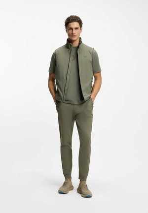 Olive green lightweight vest over a matching shirt, paired with tapered pants featuring textured details. Beige sneakers with blue accents.