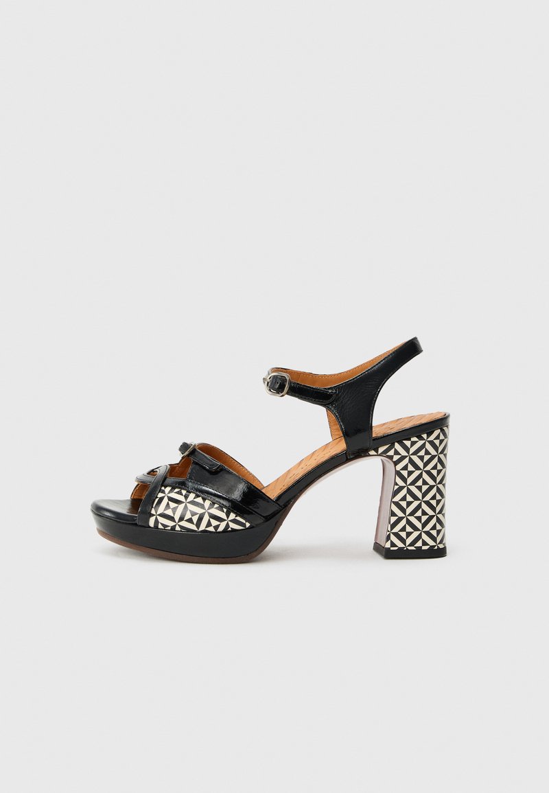 Mustast lakknahast kõrgete kontsadega sandaal koos pahkluu rihmaga, millel on geomeetrilise mustriga plokkkonts ja avatud varbaga disain.