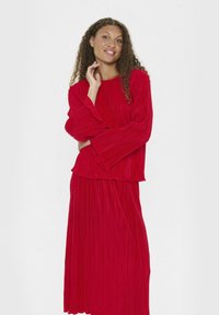 Donna con capelli ricci che indossa una maglietta rossa a maniche lunghe e una gonna rossa plissettata abbinata, sorridente e posando davanti a uno sfondo bianco.