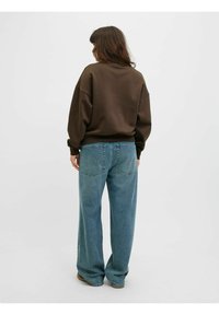 Felpa oversize marrone con spalle scese, abbinata a jeans a gamba larga blu chiaro con tasche posteriori. Sfondo semplice.