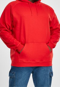 Man in een felrode hoodie met een buidelzak en blauwe cargo jeans, met zijn handen in de zakken van de hoodie, tegen een effen achtergrond.