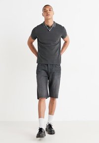 Camisa polo gris oscuro combinada con pantalones cortos de mezclilla oscuros hasta la rodilla. El modelo lleva calcetines blancos y zapatos negros, de pie frente a un fondo blanco.