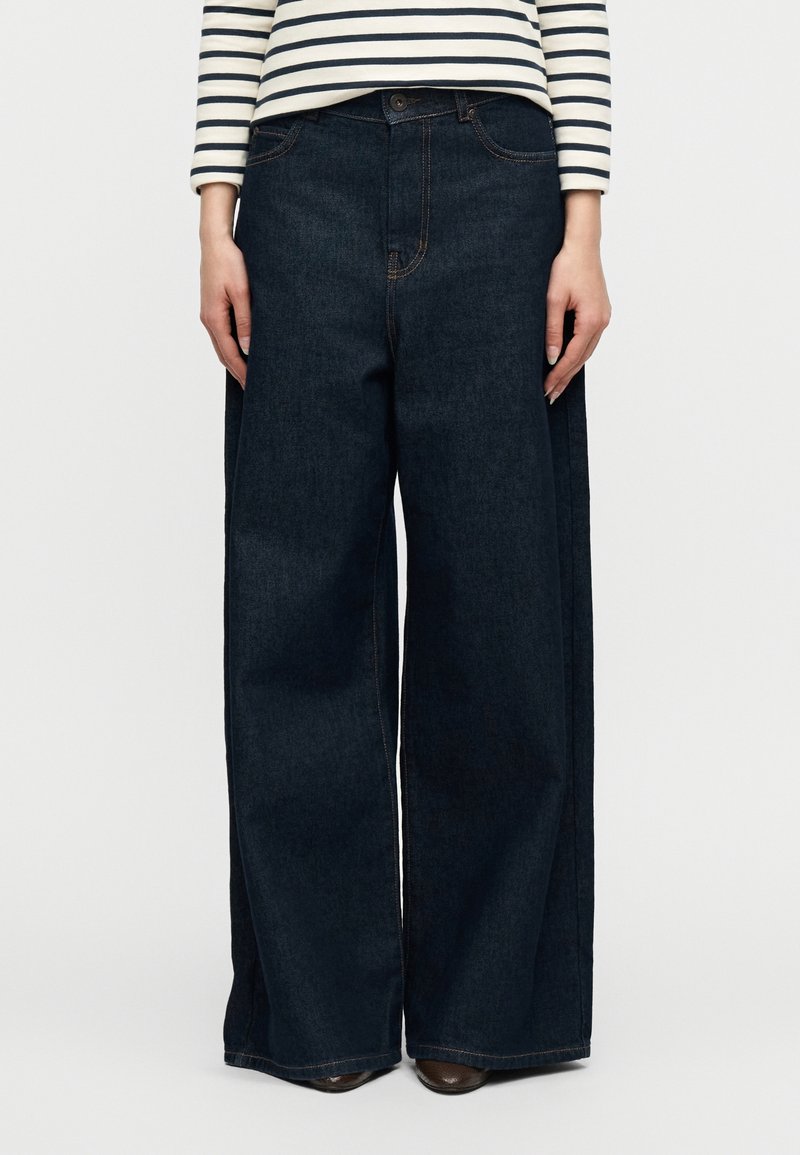 Personne portant un jean large taille haute de couleur foncée et un haut blanc à manches longues avec des rayures horizontales noires sur un fond uni.