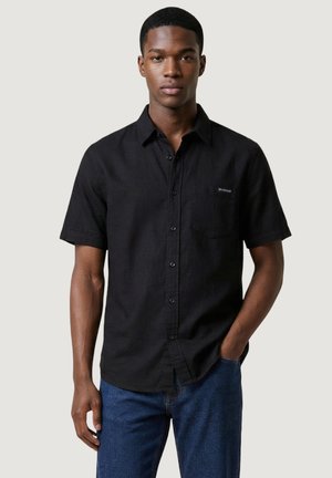 Giovane con indosso una camicia nera a maniche corte con taschino sul petto e jeans blu, in piedi con una mano in tasca.