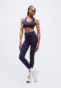 Αθλητική γυναίκα ποζάρει φορώντας μωβ αθλητικό σουτιέν και κολάν με λευκά λογότυπα Puma, λευκές κάλτσες και λευκά αθλητικά παπούτσια μπροστά σε απλό φόντο.