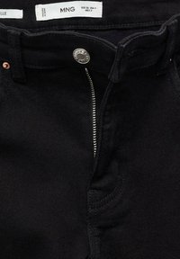 Jean noir avec fermeture éclair partiellement ouverte, bouton argenté et étiquette de taille indiquant EUR 36, USA 4, fabriqué au Pakistan.