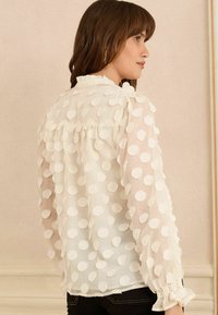 Blusa bianca in tessuto trasparente, caratterizzata da grandi pois bianchi. Presenta maniche a sbuffo e polsini arricciati. Materiale morbido e leggero.