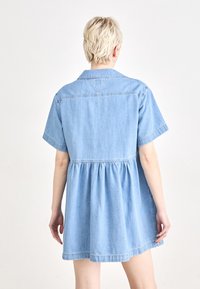 Ljusblå denimklänning med korta ärmar, krage och veckad midja, som har en utställd nederdel och minimala sömdetaljer.