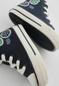 Granatowe płócienne trampki typu high-top z białymi gumowymi podeszwami, z naszytymi łatkami i kontrastującymi sznurówkami, zaokrąglonym czubkiem oraz szczegółowymi wzorami na podeszwie.