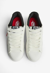 Pari valkoisia tennareita, joissa on paksut valkoiset nauhat, mustat pehmustetut nilkkakotelot ja punaiset sisäpohjat, joissa on koko 42 ja logo.