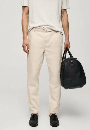 Mann trägt beigefarbene Hose, cremefarbenes Hemd, schwarze Loafers, hält schwarze Lederreisetasche an den Griffen vor einfarbigem Hintergrund.