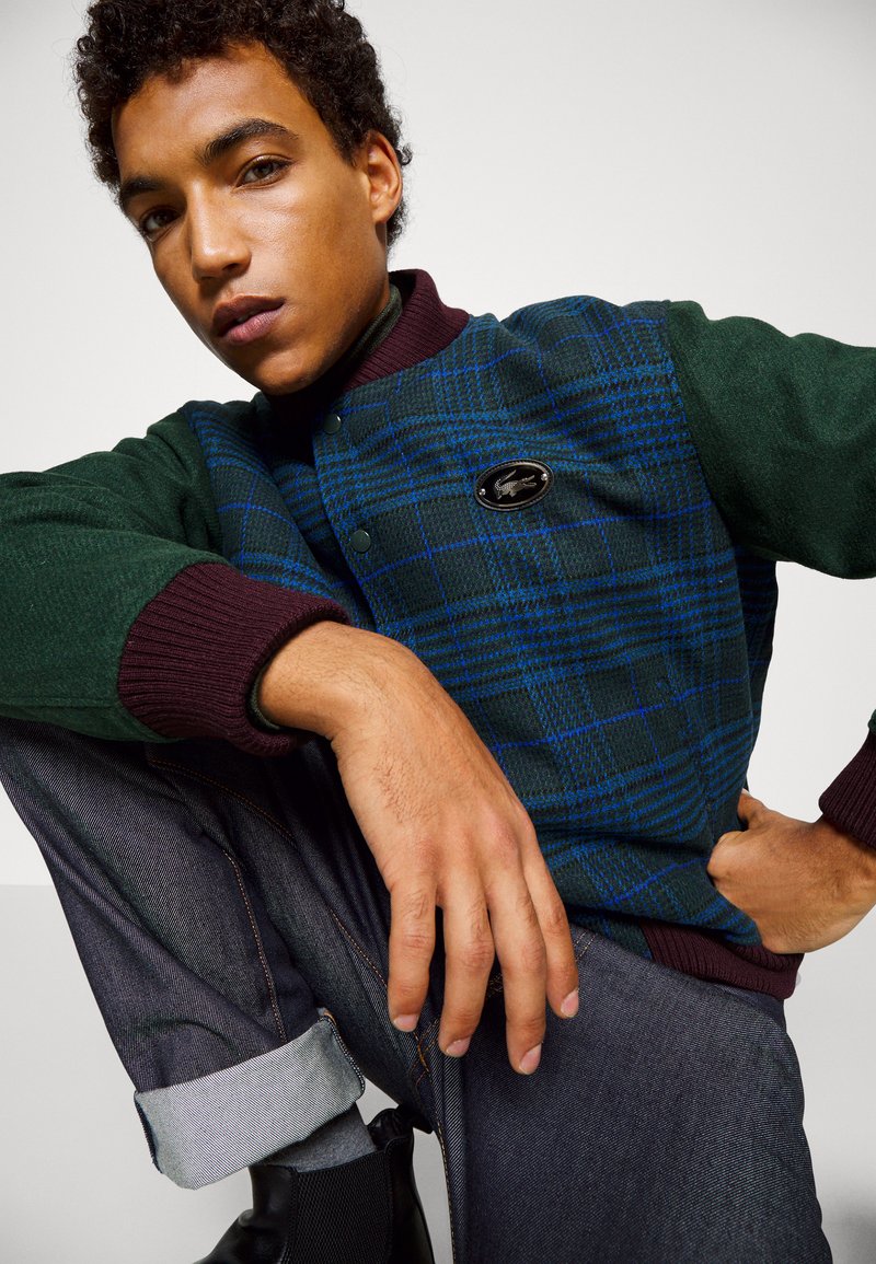 Veste bomber à carreaux verts et bleus avec des poignets côtelés bordeaux, arborant un badge logo noir. Associée à un jeans en denim foncé.