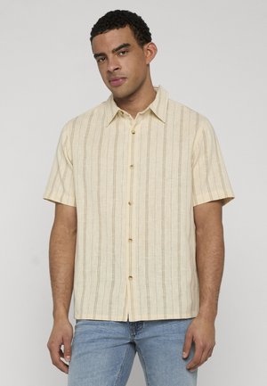 Homme portant une chemise beige à manches courtes avec des rayures verticales et un jean bleu clair, debout devant un fond gris uni.