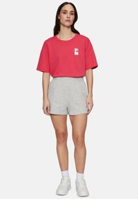 Maglietta rossa di cotone con logo quadrato, abbinata a pantaloni corti grigi chiari. Il modello indossa sneakers bianche e calzini alla caviglia. Design semplice e casual.