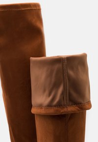 Botas hasta la rodilla de ante marrón con una sección superior suave y doblada. Incluyen detalles de costura y un acabado suave, destacando los contrastes de textura.