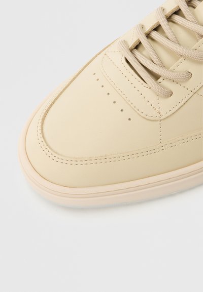 Beež värvi tossu varbakate, millel on tekstureeritud paelad, õmmeldud detailid ja väikesed augud heledal taustal.