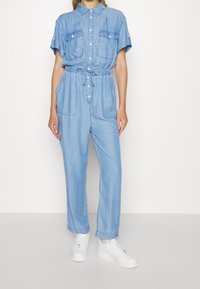 Kvinna iklädd en ljusblå kortärmad denimoverall med framfickor och dragsko i midjan, som är kombinerad med vita sneakers.