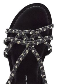 Sandale plate noire avec des courroies tressées ornées de petits embellissements brillants et une semelle intérieure lisse et mate.