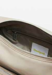 Interno di una borsa in tessuto beige con una superficie liscia, cerniera argentata e un'etichetta bianca con la scritta "Desigual" in giallo.