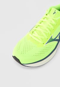 Λαμπερό κίτρινο παπούτσι για τρέξιμο με επάνω μέρος από διαπνέον πλέγμα, υφή επιφάνειας και αντίθετες μπλε πινελιές στο πλάι και στη σόλα.