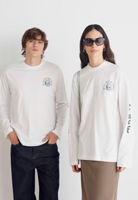 Hvit langermet t-skjorter med grafisk trykk på brystet og grønn tekst på ermene, kombinert med mørke jeans og et brunt skjørt.