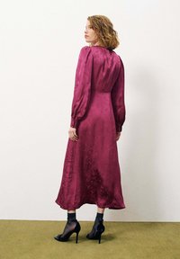 Robe longue bordeaux en tissu lisse, avec des manches bouffantes et un design au dos. Portée avec des chaussures à talons hauts et des chaussettes transparentes.