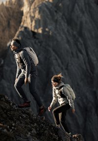 Zwei Wanderer mit Rucksäcken und Stirnbändern klettern bei schwachem Sonnenlicht einen steilen felsigen Berghang hinauf.