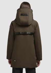 Personne portant un manteau à capuche brun avec des sangles noires de marque dans le dos, se tenant avec les mains détendues sur les côtés.