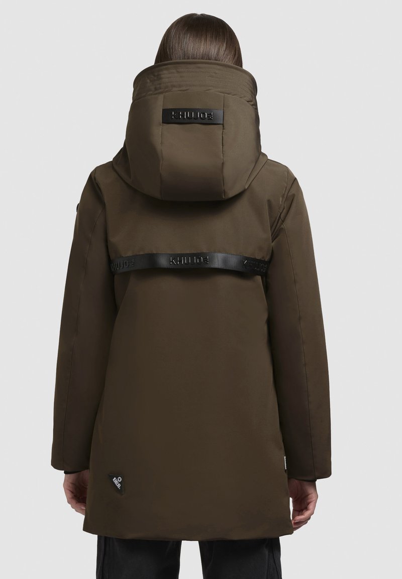 Persona vistiendo un abrigo con capucha marrón y correas negras de marca cruzando la espalda, de pie con las manos relajadas a los lados.