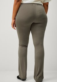 Een persoon die met de rug naar de camera staat, gekleed in olijfgroene strakke broek en zwarte schoenen, tegen een eenvoudige lichte achtergrond.