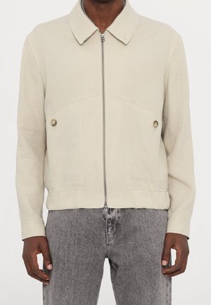 Beige jakke med lynlås, krave og knaplommer, båret med grå vaskede jeans på en stående person.