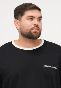 Zwart T-shirt met korte mouwen en een crèmekleurige ronde hals, voorzien van witte geborduurde tekst "Originals Studio" op de linkerkant van de borst.