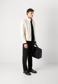 Man wearing white zip-up jacket and black pants holding black leather bag, standing against plain white background.