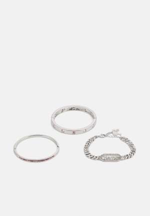 Trois bracelets en métal : un fin en argent avec des pierres roses, un plus large avec des pierres claires et roses alternées, et un bracelet en chaîne épais avec un logo.