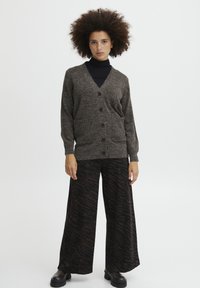 Cardigan marrone lavorato a maglia con bottoni, dotato di due tasche frontali; abbinato a pantaloni larghi neri con stampa zebra scura e stivali neri robusti.