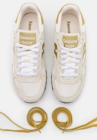 Paar beige en gouden Saucony Shadow sneakers met witte veters geknoopt, vergezeld door aparte opgerolde goudkleurige veters op een witte achtergrond.